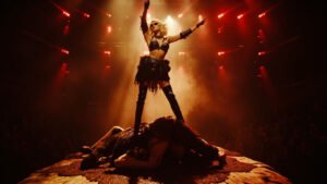 Lady Gaga standing victorious over a fallen dancer on a dark theatrical stage with fiery backdrop during her Mayhem Ball tour performance